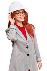Young redhead woman wearing architect hardhat smiling with hand over ear listening an hearing to rumor or gossip. deafness concept.