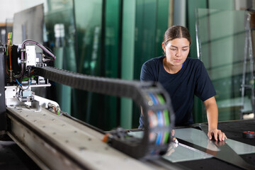 Young workwoman using manual glass cutter to precisely cut sheet of glass in processing workshop...