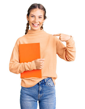 Beautiful caucasian woman with blonde hair holding book pointing finger to one self smiling happy and proud