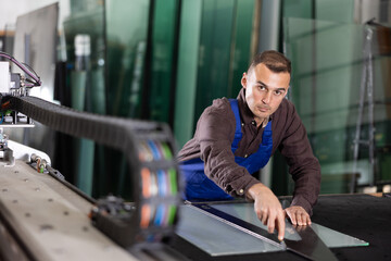 Focused male technician in work shirt and blue overalls meticulously cutting glass sheet with...