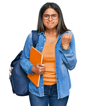 Young latin girl wearing student backpack and holding books angry and mad raising fist frustrated and furious while shouting with anger. rage and aggressive concept.