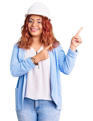 Young latin woman wearing architect hardhat smiling and looking at the camera pointing with two hands and fingers to the side.