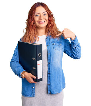 Young latin woman holding file cabinet pointing finger to one self smiling happy and proud