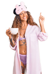 Young hispanic woman with tattoo wearing sleep mask and pajama drinking coffee surprised with an...