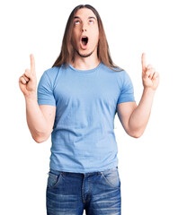 Young adult man with long hair wearing casual clothes amazed and surprised looking up and pointing with fingers and raised arms.