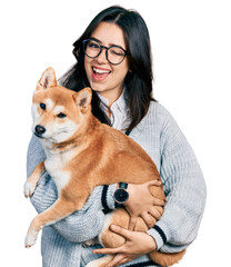 Beautiful hispanic woman holding cute dog winking looking at the camera with sexy expression, cheerful and happy face.
