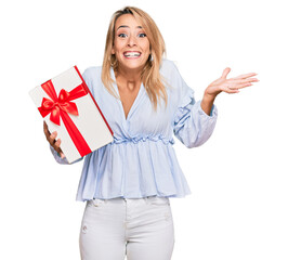 Beautiful blonde woman holding gift celebrating victory with happy smile and winner expression with raised hands