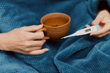 Person holding cup of coffee and thermometer, sitting on blanket with another blanket over them
