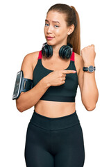 Beautiful blonde woman wearing gym clothes and using headphones in hurry pointing to watch time, impatience, looking at the camera with relaxed expression
