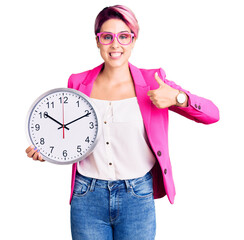 Young beautiful woman with pink hair wearing business jacket and holding clock smiling happy and...