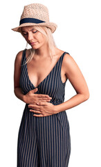 Young beautiful blonde woman wearing summer hat and dress with hand on stomach because indigestion, painful illness feeling unwell. ache concept.