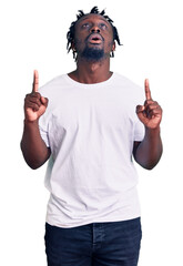 Young african american man with braids wearing casual white tshirt amazed and surprised looking up and pointing with fingers and raised arms.
