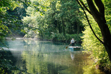 An elderly man rowing a small boat on a serene, tree-lined river in the Netherlands, surrounded by lush greenery and dappled sunlight, creating a peaceful, idyllic scene