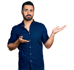 Young hispanic man with beard wearing casual blue shirt clueless and confused expression with arms...