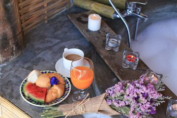 Croissant, watermelon with hot coffee and orange juice near bathtub with candle on wooden plank. Relaxation on holiday vacation.