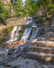 Stairs leading up a beautiful cascading waterfall surrounded by lush greenery. Cascadilla Gorge in Ithaca New York- Finger Lakes Region
