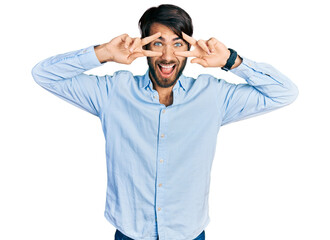 Fototapeta premium Hispanic man with blue eyes wearing business shirt doing peace symbol with fingers over face, smiling cheerful showing victory