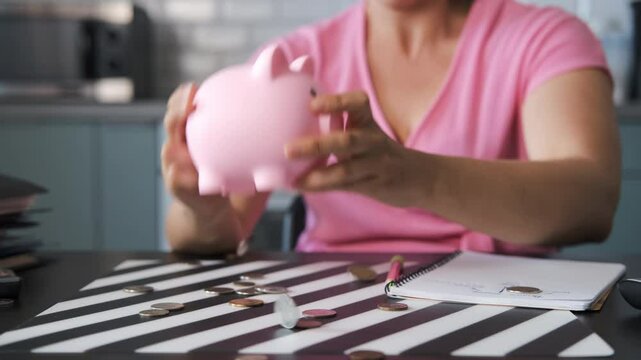 Close-up of young woman empties a piggy bank, coins falling and scattering on a kitchen table. Concept of budgeting and managing personal finances