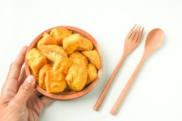 Someone hand holding and serving fried yellow tofu on wooden plate next to wooden spoon and fork. Tahu Takwa is a specialty food from Kediri, East Java, Indonesia. Flat Lay Photo