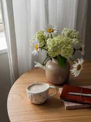 Coffee break - a cup of cappuccino, a vase with flowers, a notebook, a book on a round wooden table