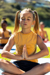 Naklejka premium Composite of caucasian schoolgirl meditating in prayer pose while sitting on mat in park