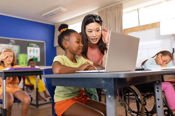Asian female teacher assisting african american schoolgirl in using laptop at desk in classroom