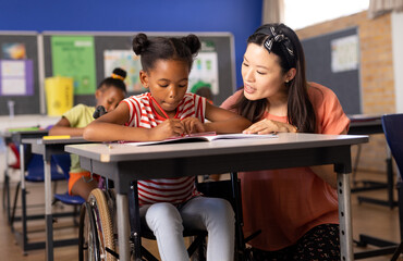 Asian female teacher assisting african american schoolgirl sitting on wheelchair to write notes