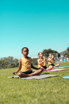 Fototapeta Composite of diverse schoolgirls meditating while sitting on mats over grassy field under clear sky