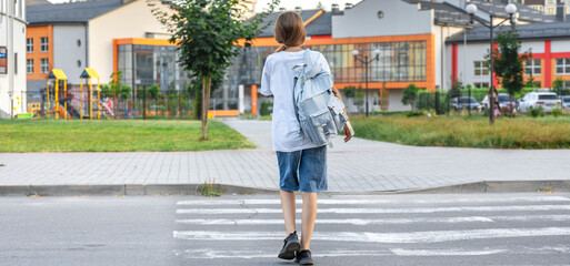 A schoolgirl crosses a pedestrian crossing. Background of the school. Back view. Goes to school.