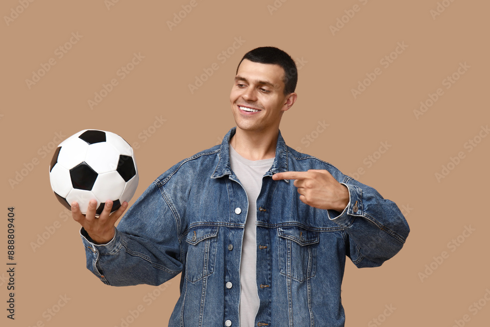 Wall mural handsome young man pointing at soccer ball on brown background