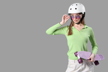 Beautiful young woman in helmet with skateboard on grey background