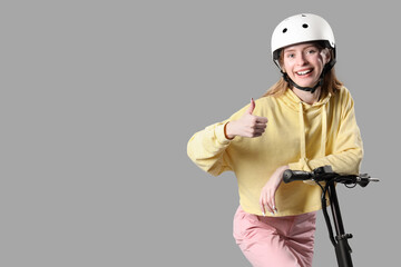 Beautiful young woman in helmet with modern electric kick scooter showing thumb-up gesture on grey background