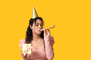 Beautiful young African-American woman in party hat with whistle and gift box on yellow background