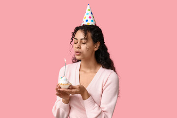 Beautiful young African-American woman in party hat holding birthday cupcake with burning candle on...