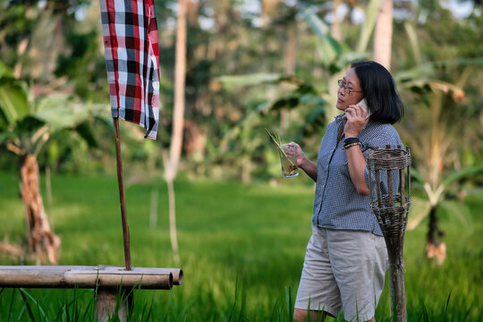 Woman standing and answering the phone while other hand holding a glass of cold iced tea in the rice field