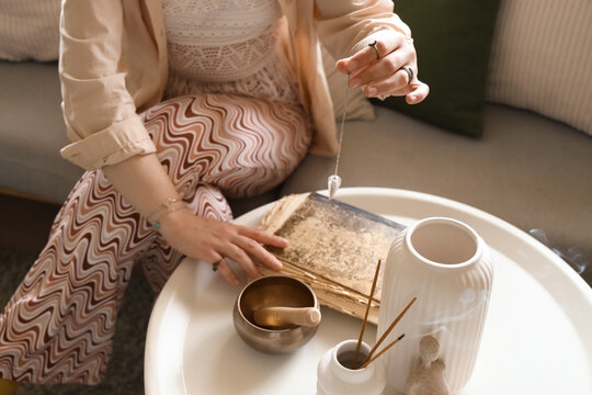 Young witch with pendulum and spell book on table at home, closeup