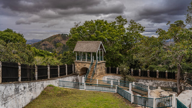 An ancient palace. The courtyard. A stone staircase rises to a gazebo with a thatched roof. Perimeter fence. Lush tropical vegetation. Cloudy sky. Madagascar. Royal Hill of Ambohimanga. Antananarivo.