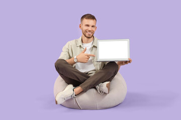 Handsome man pointing at blank laptop on lilac background