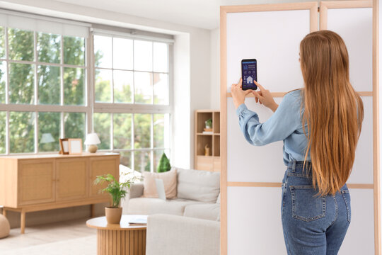 Young woman using mobile phone with smart home security system control panel in room, back view