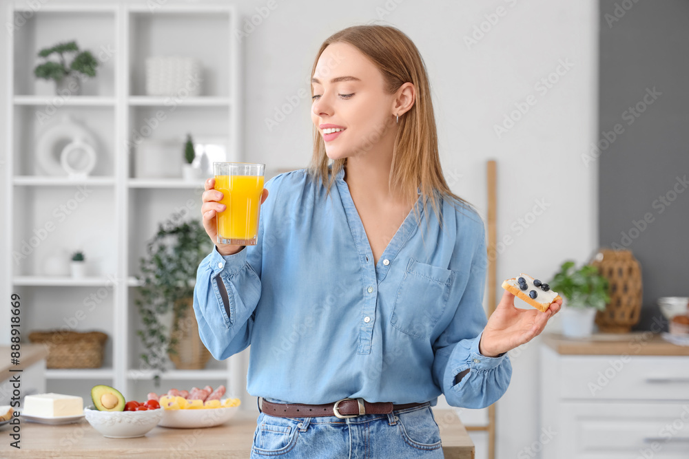 Poster Beautiful young happy woman holding glass of juice, tasty toasts with butter and blueberries at home