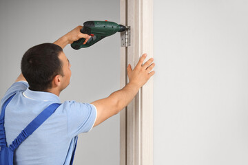 Male worker with electric drill installing door hinge at home
