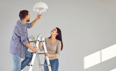 Young man standing on the ladder changing light bulb in the room with his smiling wife. Happy...