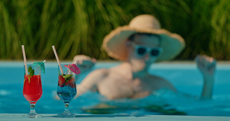 Funny man in a straw hat dancing joyfully in the pool with two cocktails on the edge