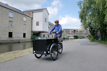 White middle aged man rides a large black electric cargo bike along a canal towpath in the summer.