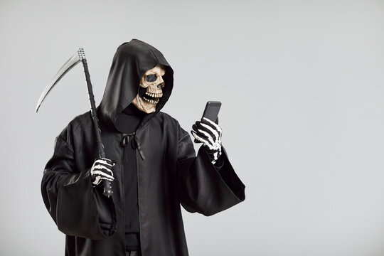 Awful grim reaper, with a scythe and wearing skeleton mask, uses a mobile phone on a grey background. This eerie image combines the reaper deathly presence with online, internet elements.