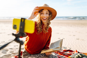 A young woman sits on the beach and records a video for her subscribers. Happy woman creates content. Travel, blogging, technology concept