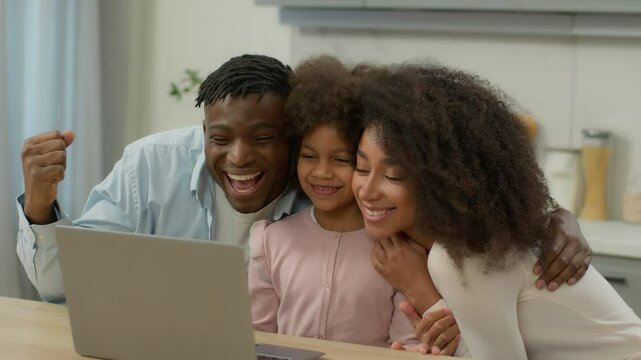 Happy family African American mother father with little daughter win laptop using pc celebrate achieve food delivery order discount dad mom with child kid baby girl computer victory at kitchen home