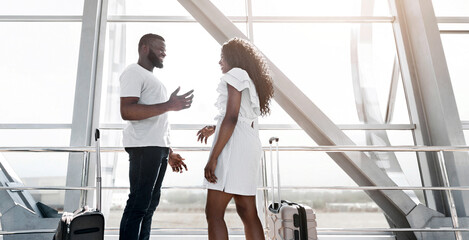 African American man and a woman stand in the airport, engaged in conversation. They are waiting to board a flight, with luggage beside them, copy space