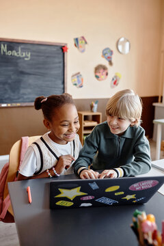 Vertical shot of two smiling primary students of black and white ethnic group studying together on laptop having fun in class, copy space