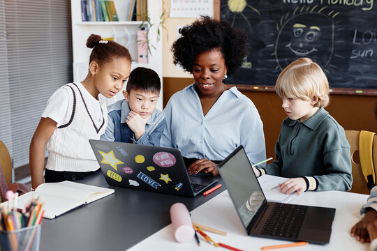 Diverse group of primary learners around female teacher of Black ethnicity explaining new information using laptop sitting at desk during elective lesson